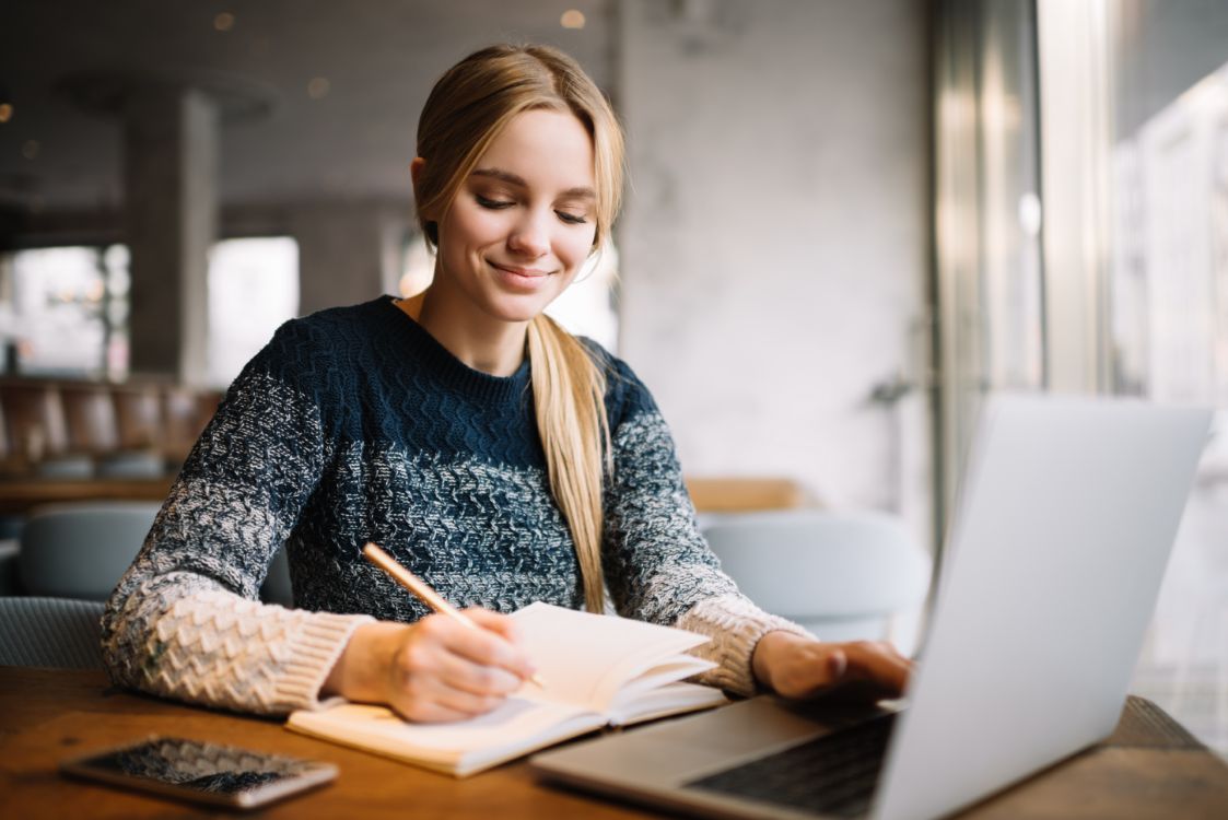 Female student  writing in notebook and working at laptop.